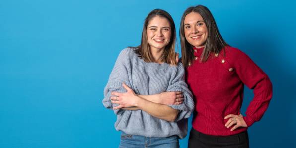 Deux jeunes femmes sourient à la caméra - celle de gauche porte un pull bleu clair, celle de droite un pull rouge à boutons dorés