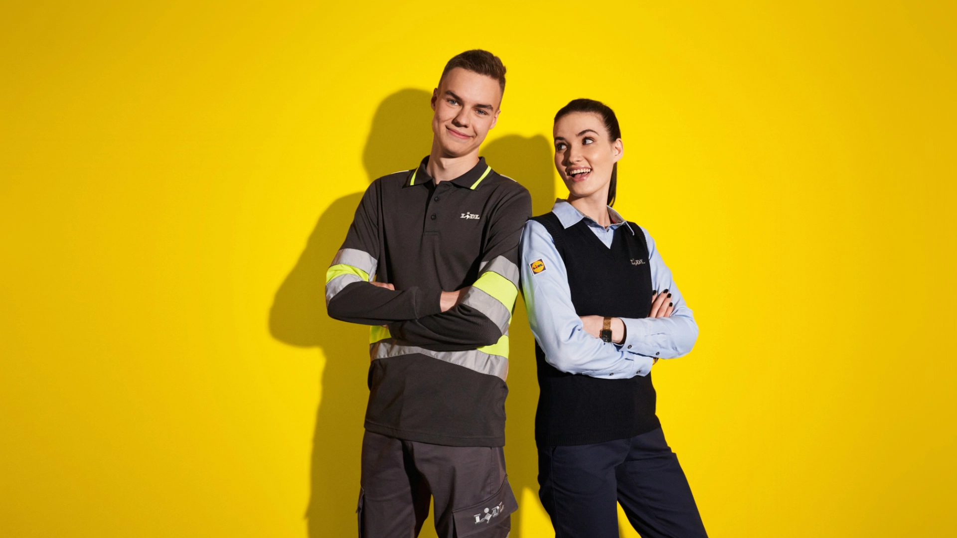Jeune homme travaillant en logistique et jeune femme en supermarché, côte à côté et complices, sur un fond jaune