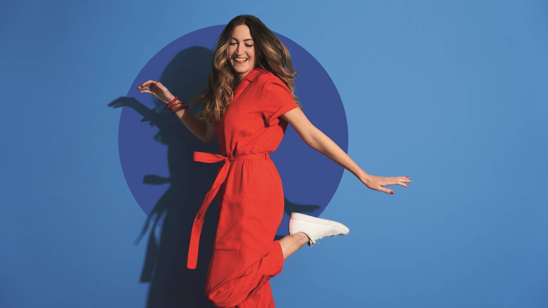 Jeune femme en combinaison rouge saute en souriant, devant un fond de couleur bleu