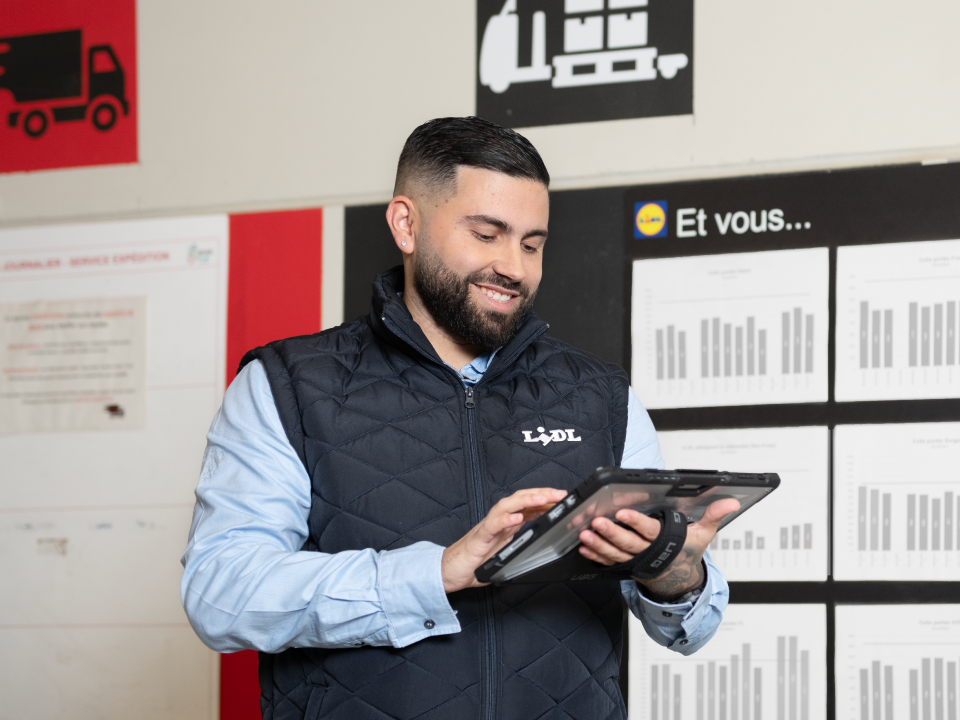Un jeune homme Responsable d'équipe logistique regarde une tablette numérique qu'il tient à la main, il se tient devant un panneau d'affichage à destination des employés