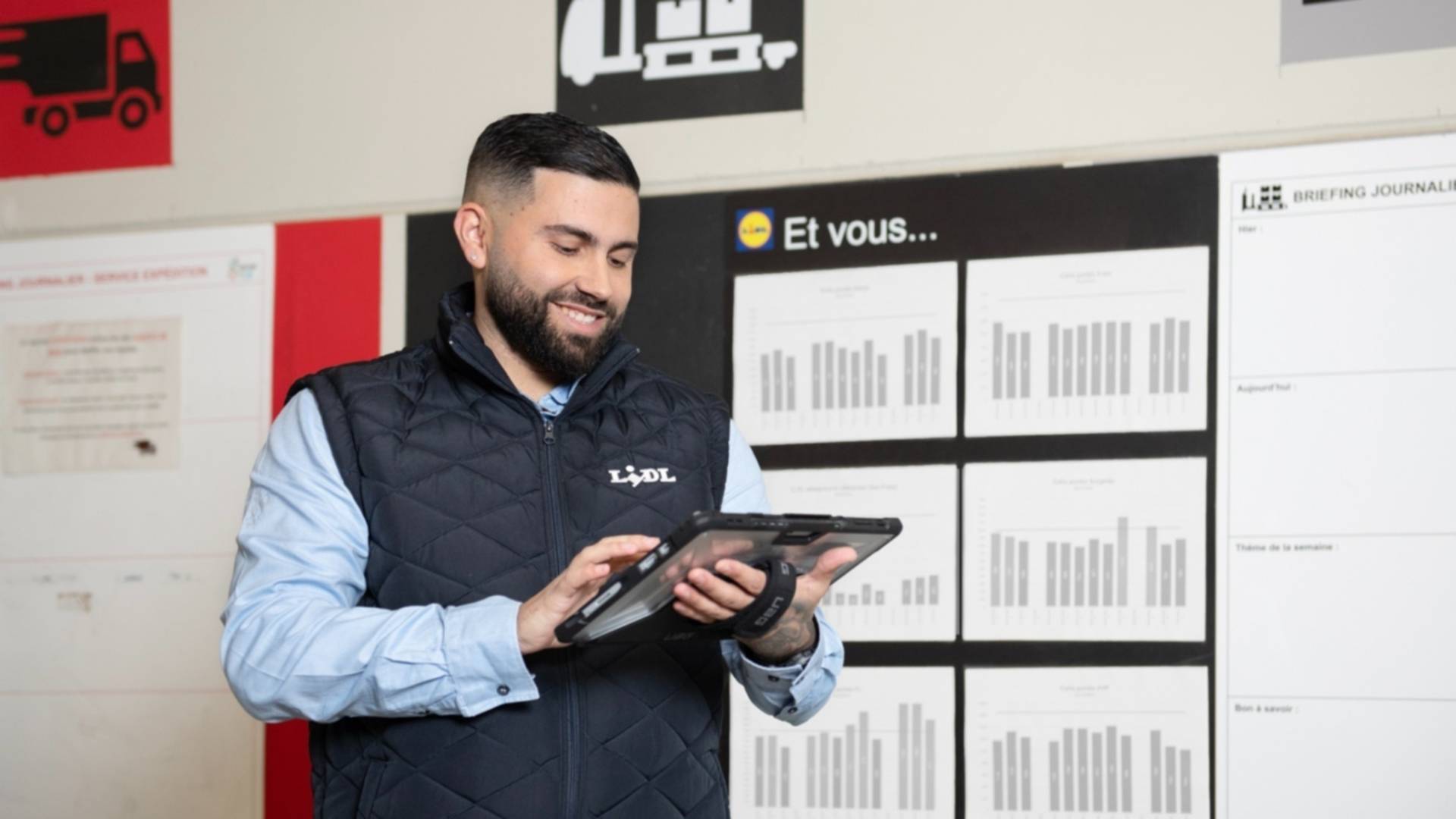 Un jeune homme Responsable d'équipe logistique regarde une tablette numérique qu'il tient à la main, il se tient devant un panneau d'affichage à destination des employés