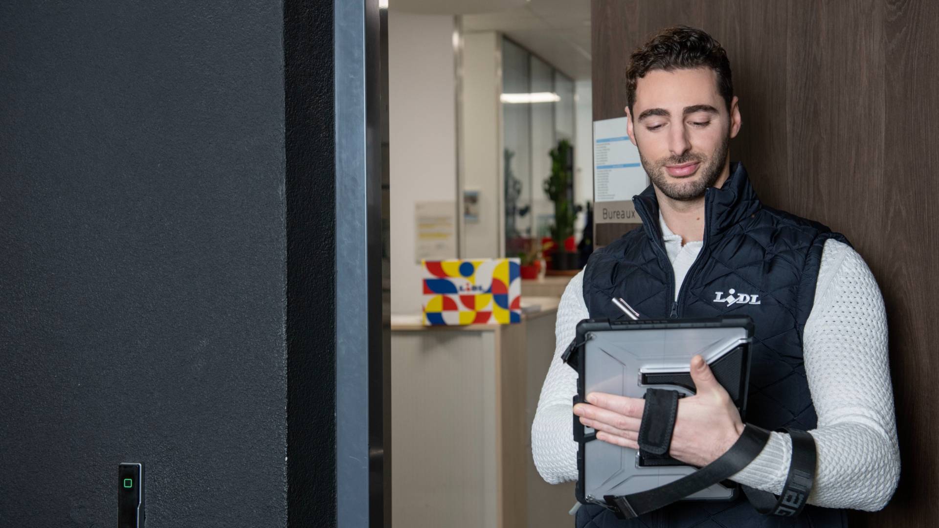 Un jeune homme, portant une doudoune Lidl noire sans manche, consulte sa tablette numérique en entrant dans son bureau