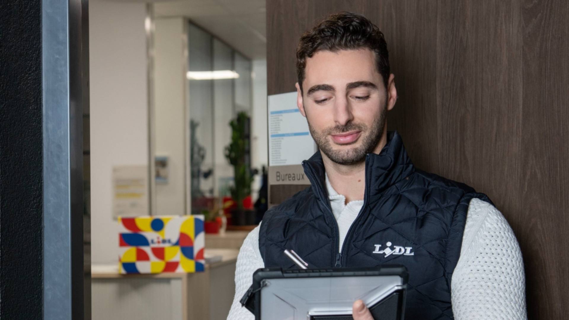 Un jeune homme, portant une doudoune Lidl noire sans manche, consulte sa tablette numérique en entrant dans son bureau